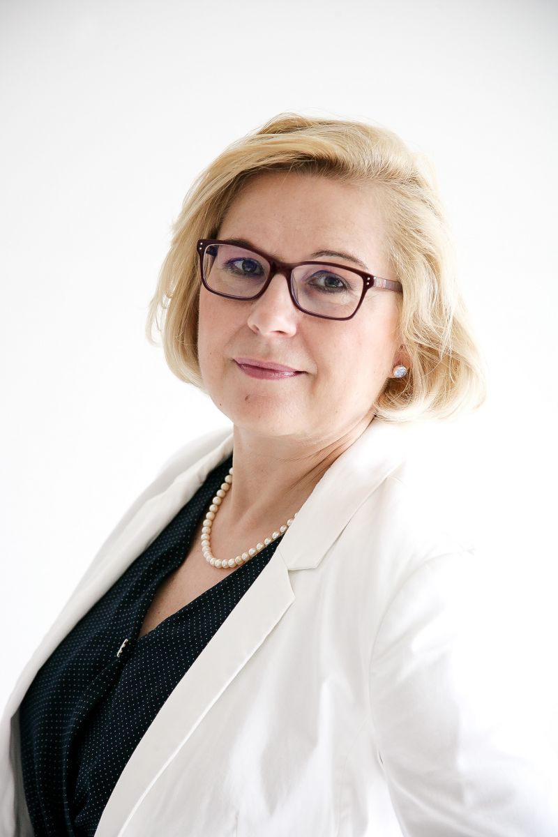 freundliche Frau, im mittleren Alter, mit blonder Bob-Frisur und Brille trägt weißen Blazer mit schwarzer Bluse und Perlenschmuck