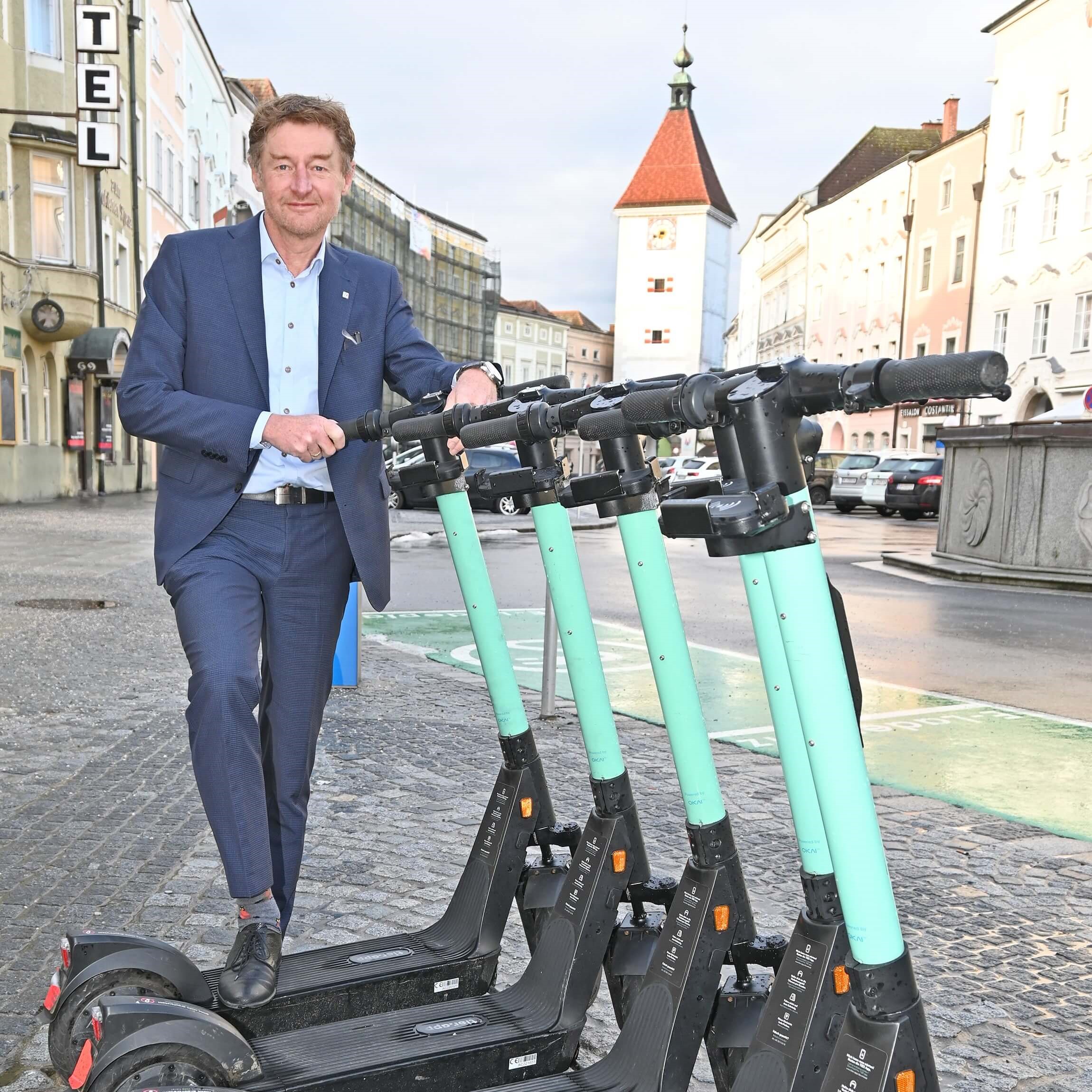Vizebürgermeister Gerhard Kroiß steht hinter einer Gruppe von E-Scootern auf dem Stadtplatz, im Hintergrund ist der Ledererturm zu sehen