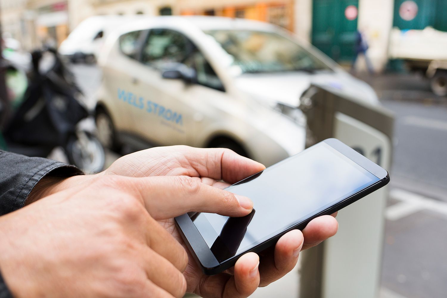 Eine Hand die auf ein Handy tippt und im Hintergrund ist ein E-Auto