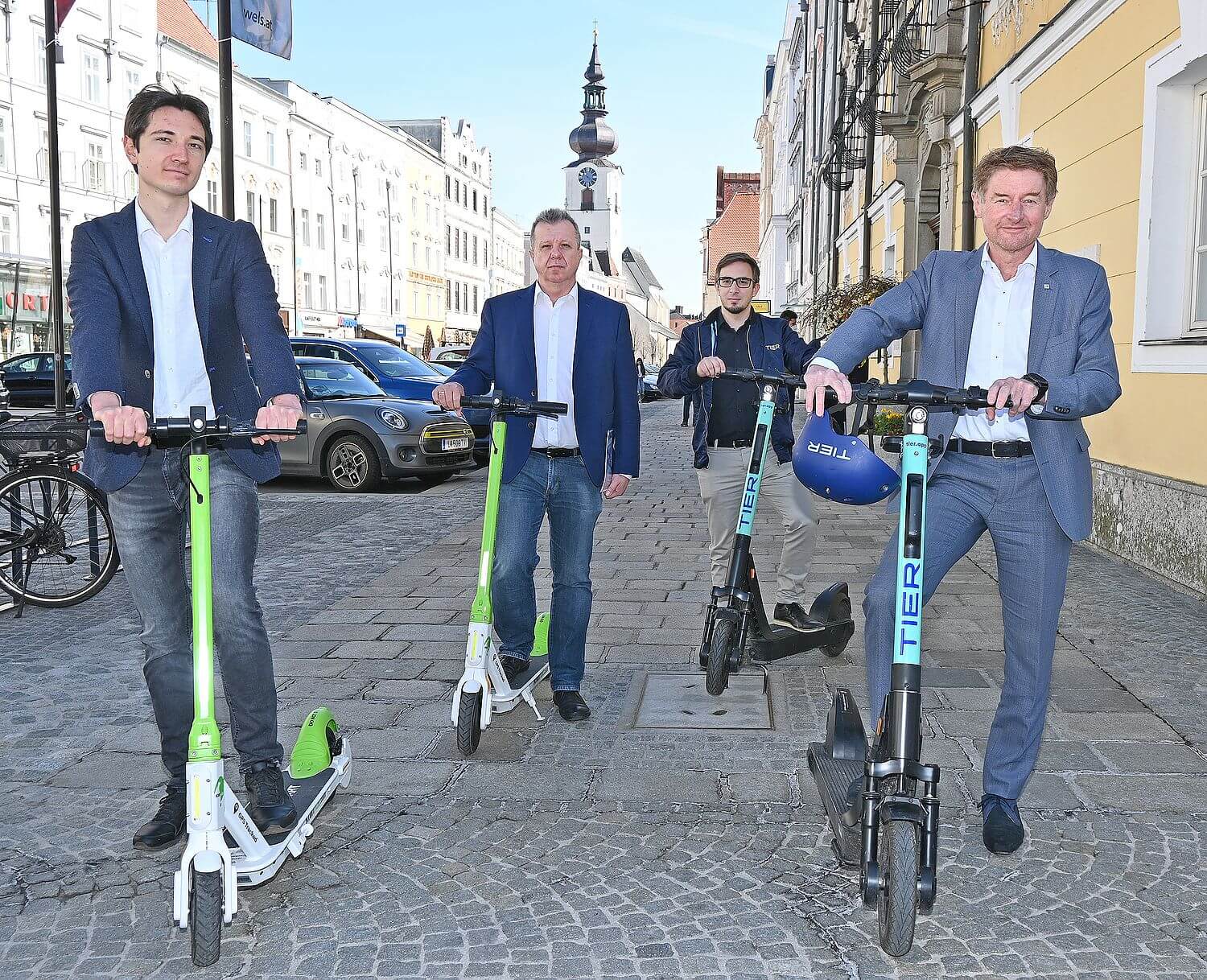 vizebürgermeister Gerhard Kroiß steht gemeinsam mit zwei Scooter Anbietern und dem Verkehrsstadtrat der Stadt Wels vor dem Rathaus. Alle Personen stehen auf einem E-Scooter