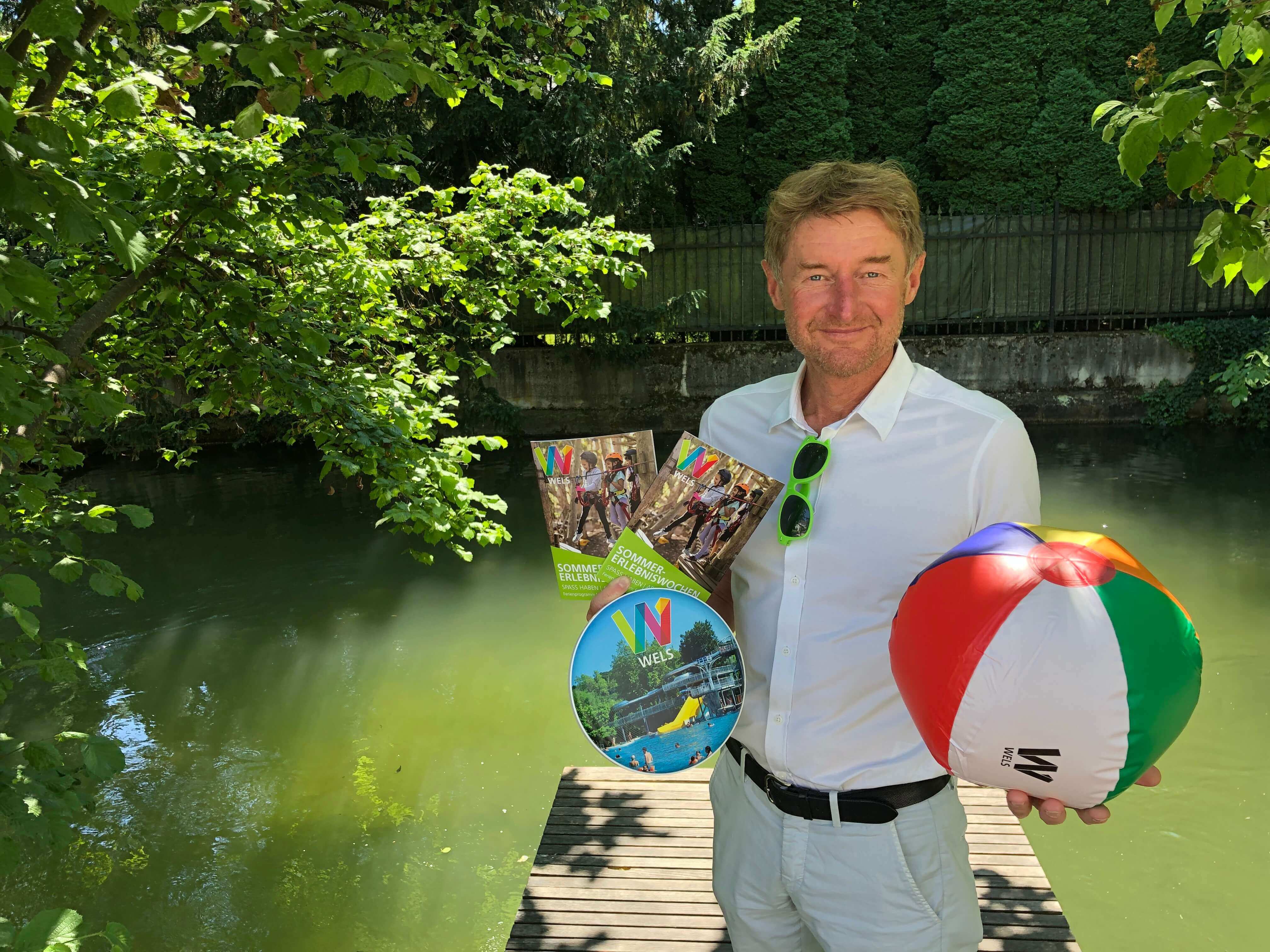 Vzbgm. Gerhard Kroiß mit einem Wasserball, Frisbee und dem diesjäjrigen Sommerferienprogramm auf einem Steg beim Welser Mühlbach