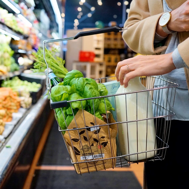 Einkaufskörbchen mit Lebensmittel, gehalten von einer Frau, im Hintergrund ein Supermarktregal